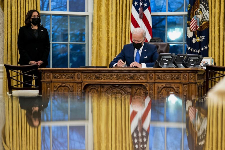 President Biden signing at a table with Kamala Harris on the left in the Oval Office