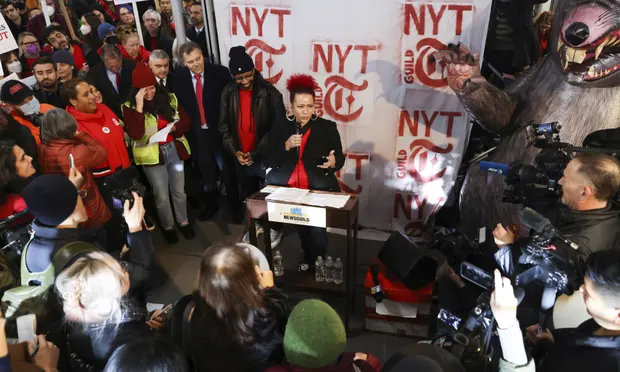 A woman with bright red hair appears speaking in front of a crowd of strikers, with red spray-paint stencils of the New York Times logo behind her.
