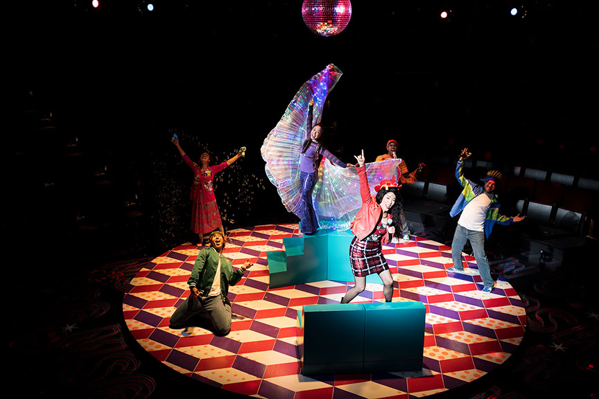 Ami stands center stage raised on green stairs, with iridescent wings. The other cast members surround her, singing, while a disco ball spins overhead and bubbles blow out of a machine.